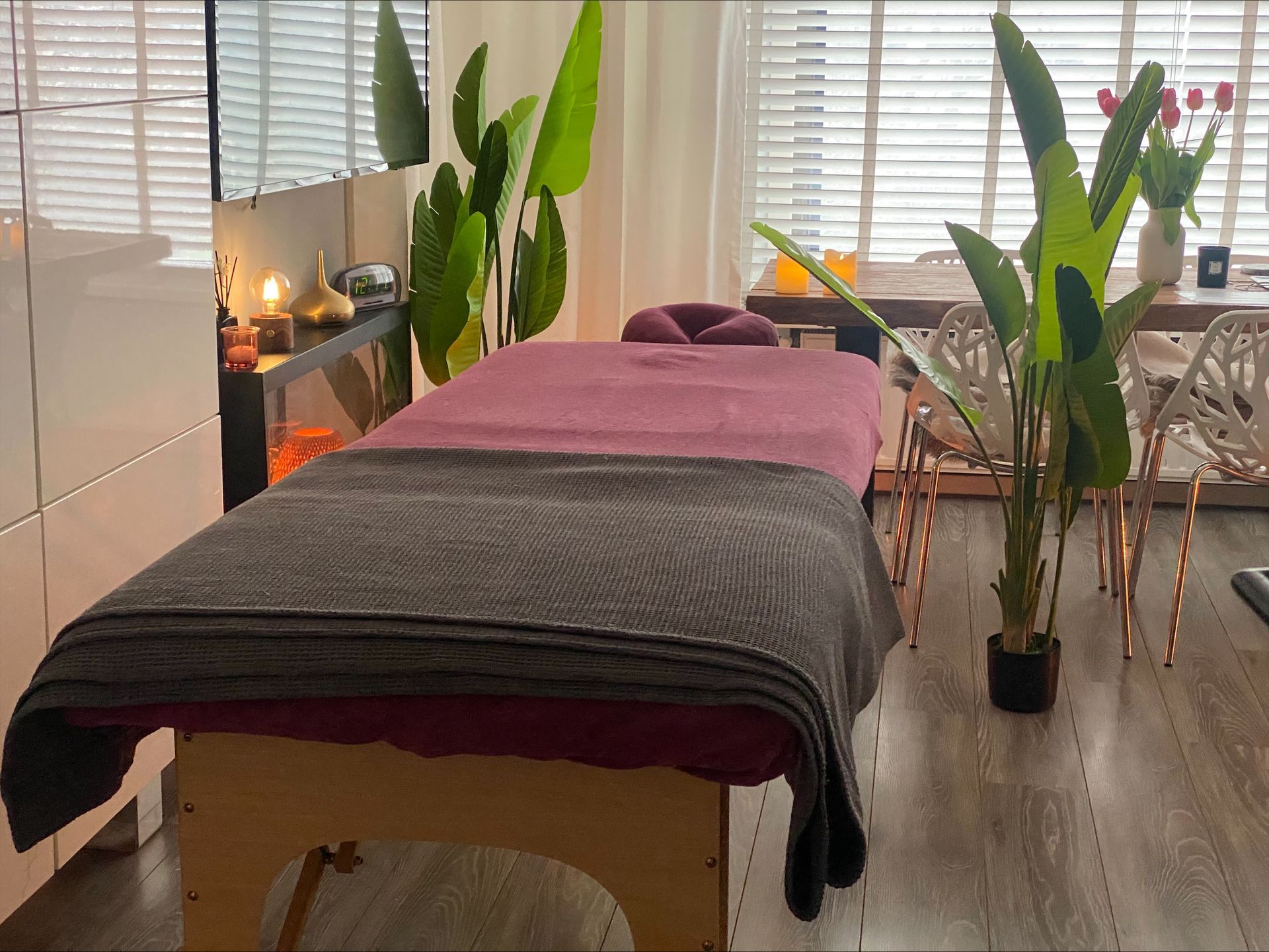Massagetafel in een lichte kamer met kaarsen, planten en een raam.