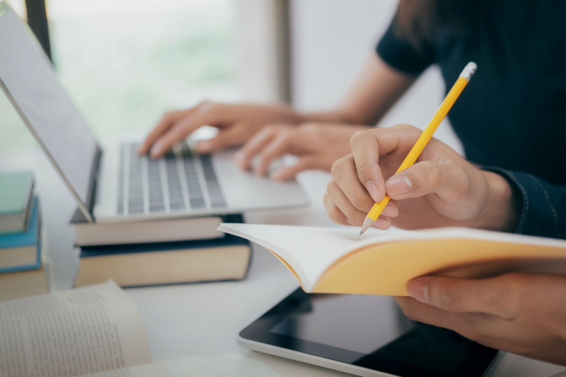 Two people writing in a notebook and typing on a laptop.