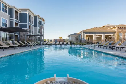 A rectangular swimming pool at an apartment complex, with lounge chairs and buildings on either side under a clear sky.