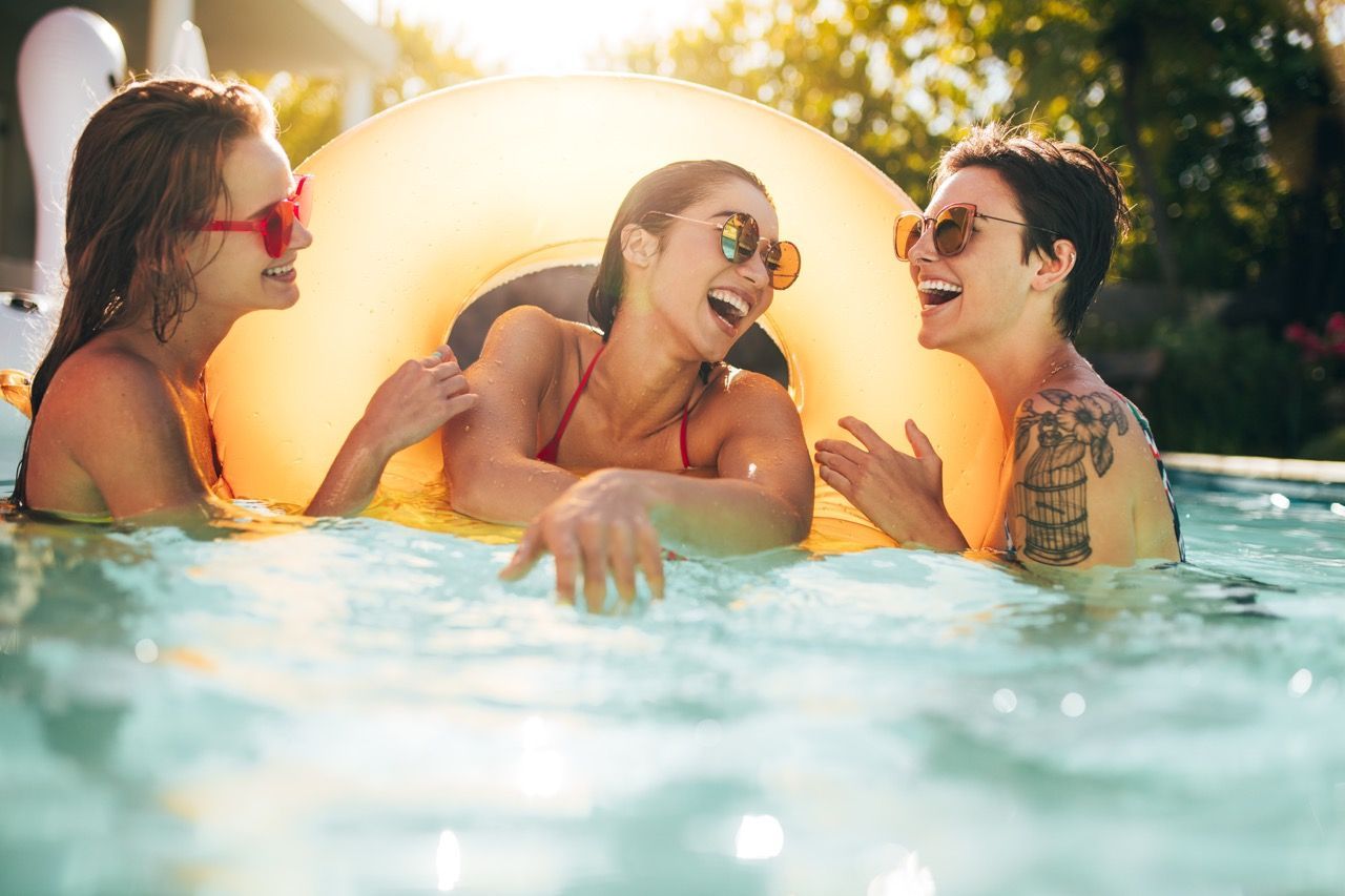 Three friends wearing sunglasses laugh while relaxing in a sunlit outdoor swimming pool with a yellow pool float.