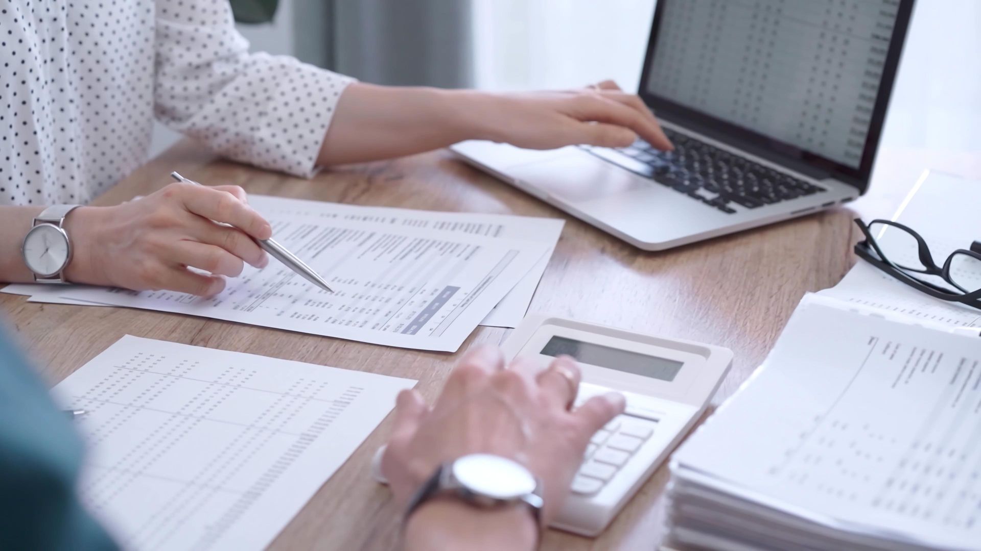 Two people work together at a wooden desk with a laptop, calculator, and financial documents. Two people work together at a wooden desk with a laptop, calculator, and financial documents.