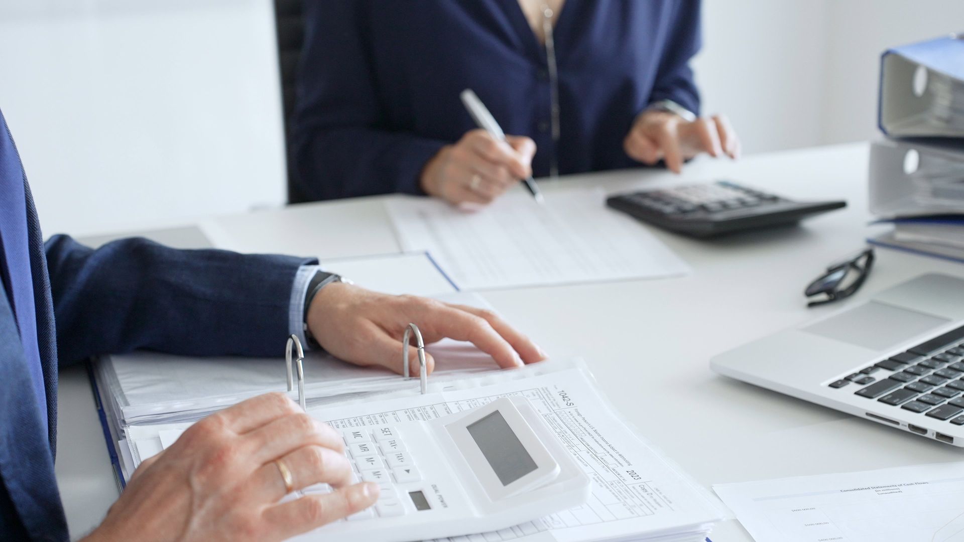 Two people in professional attire work together at an office desk, using calculators and reviewing documents. Two people in professional attire work together at an office desk, using calculators and reviewing documents.