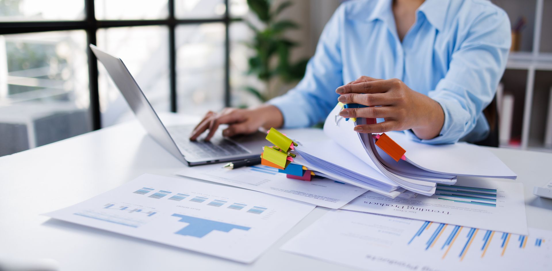 A person in a blue shirt works at a desk with a laptop and documents featuring charts and colorful binder clips. A person in a blue shirt works at a desk with a laptop and documents featuring charts and colorful binder clips.