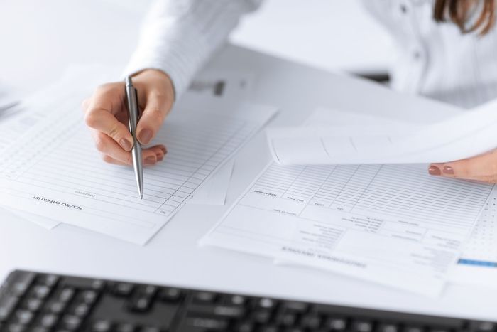 A person at a desk uses a pen to review and check items on a document filled with rows of data.