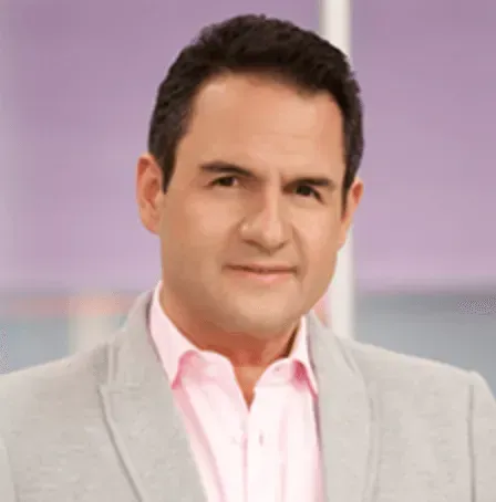 Un hombre con traje y camisa rosa está parado frente a una pared violeta.