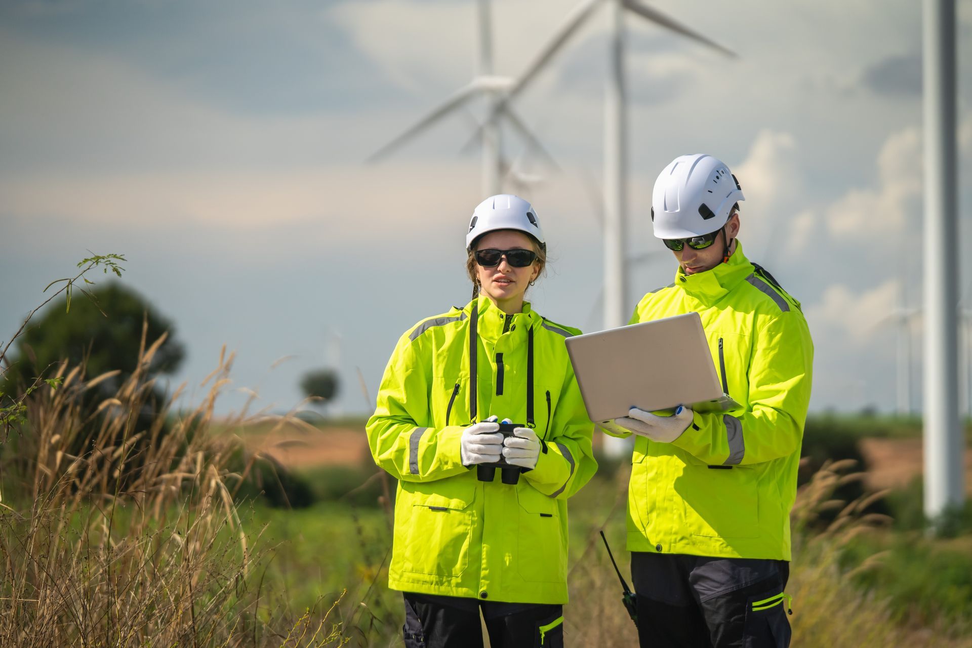 Two wind farm technicians in neon jackets and hard hats reviewing a tablet outdoors.