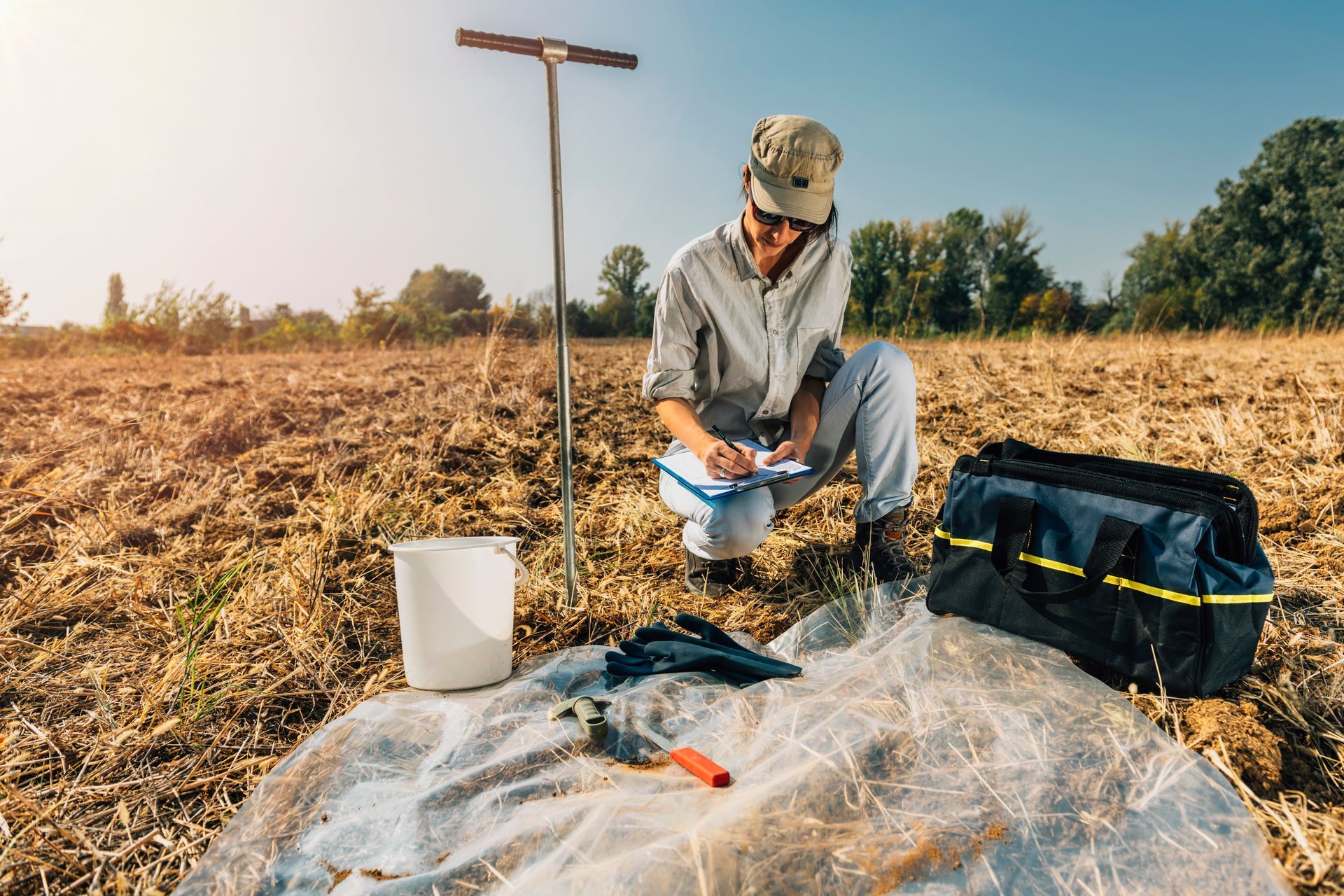 Person kneeling in a dry field, collecting soil samples beside a shovel and gear bag under clear sky