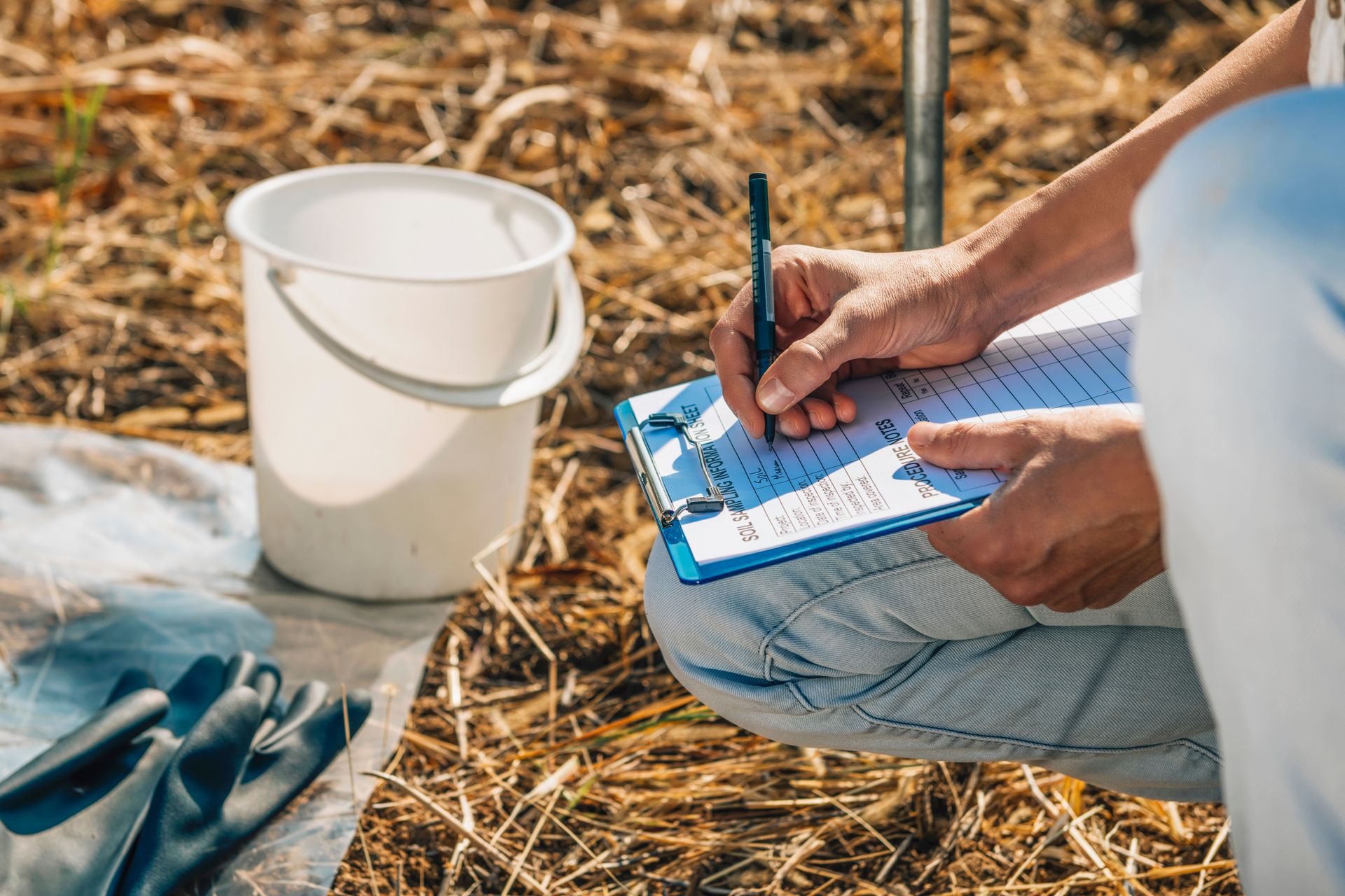 Person writing notes on a clipboard beside a white bucket outdoors on dry grass