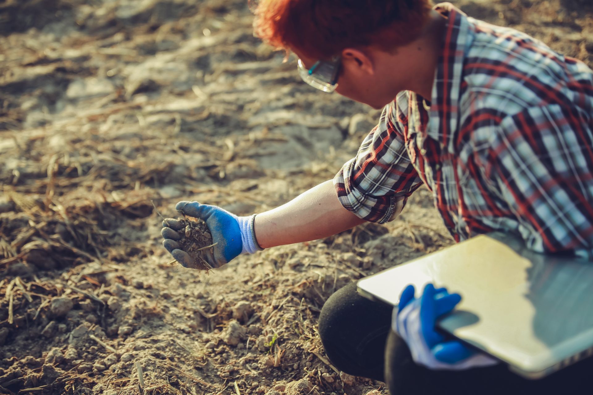 Person in plaid shirt kneeling in soil and examining a handful of dirt with gloves outdoors