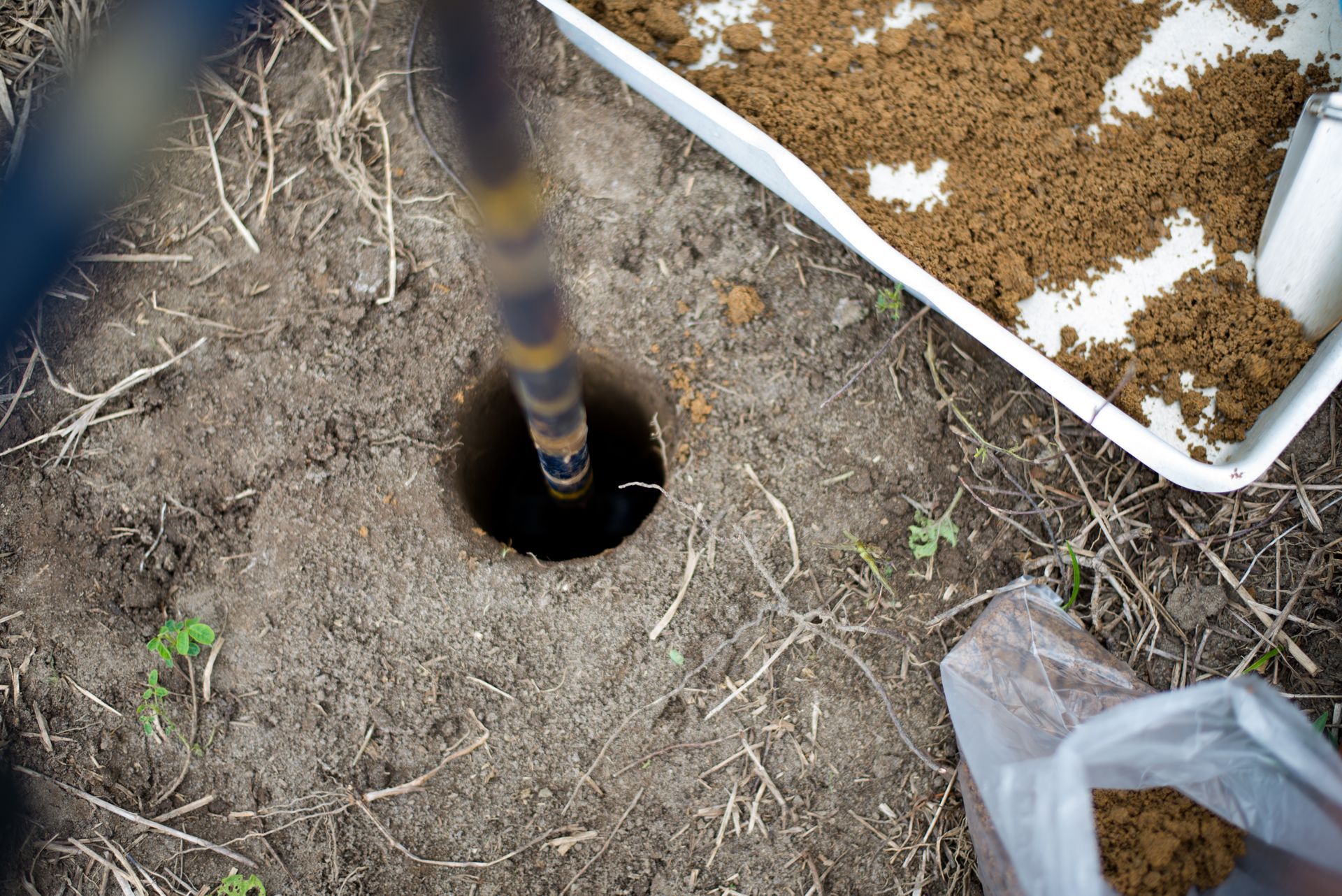 A striped pole is inserted into a round hole in the ground beside a white tray of soil and a plastic bag.
