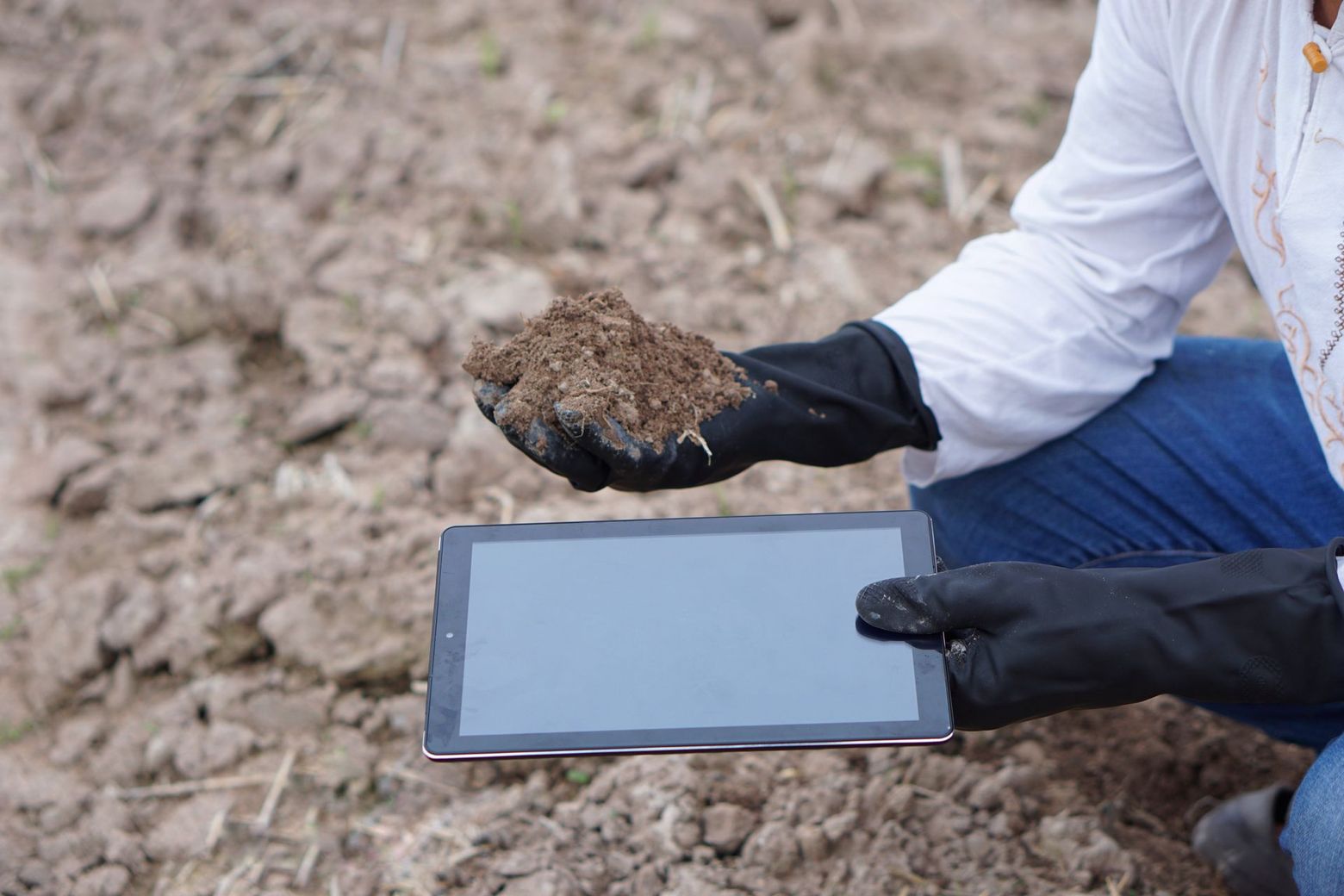 Gloved person holding soil above a tablet on dry ground, suggesting field soil analysis