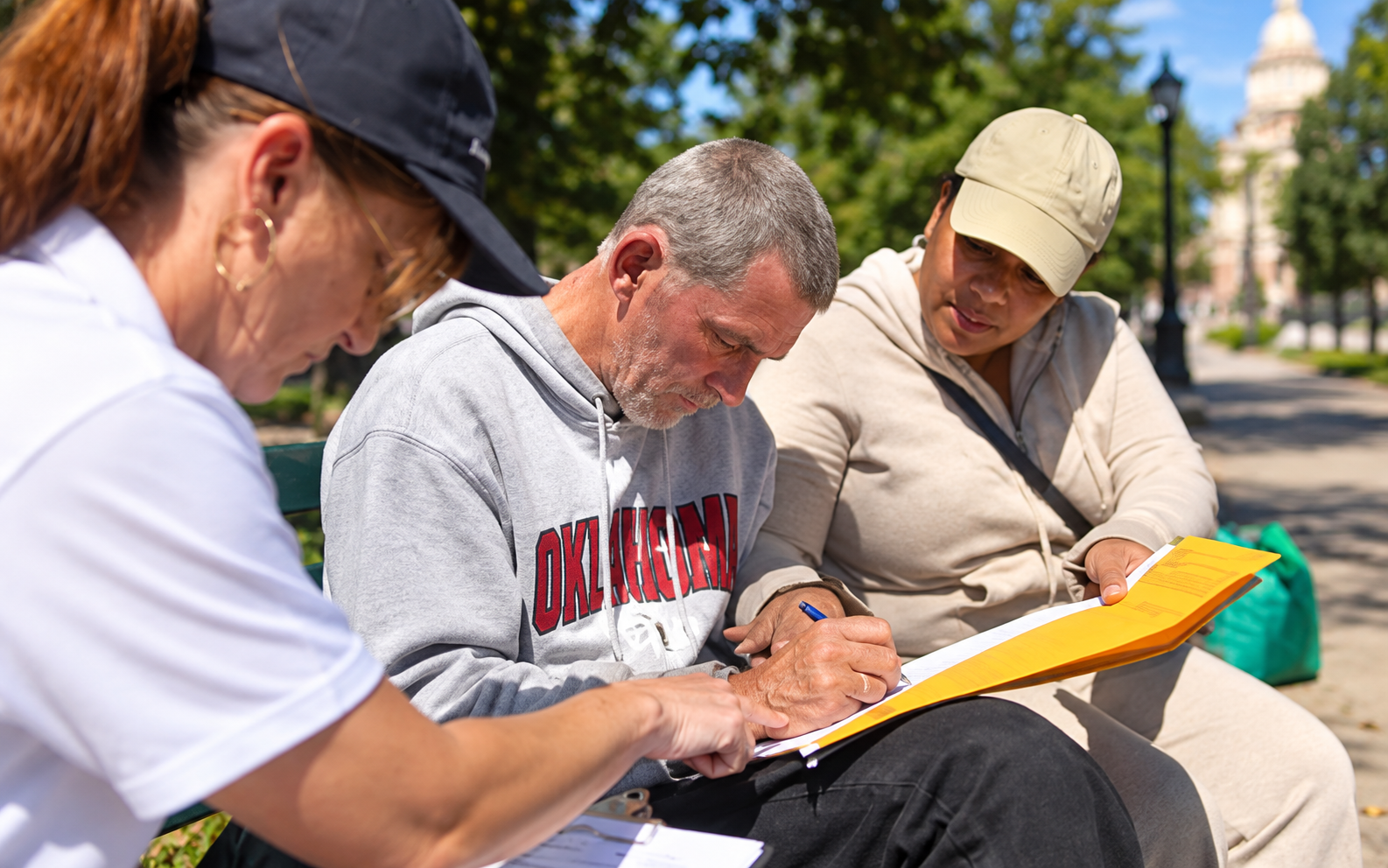 Three people sit on a bench, one writing on a clipboard, another observing, with trees and a building in the background.
