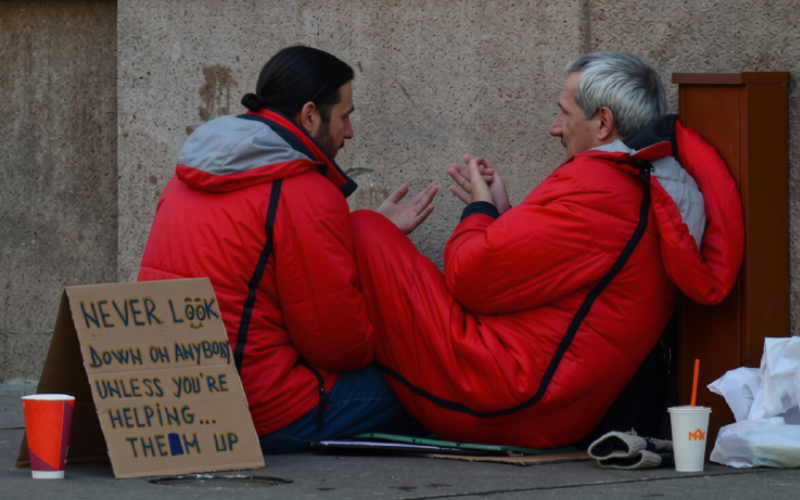 Two men in red coats sit on the sidewalk, conversing. A sign reads, "Never look down on anybody unless you're helping them up.