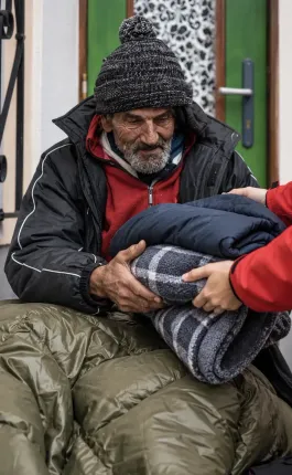 Person wrapped in a sleeping bag being assisted at a doorway, wearing a hooded jacket and red shirt