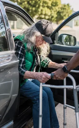 Person seated in a walker at an open car door, being assisted by another person outdoors