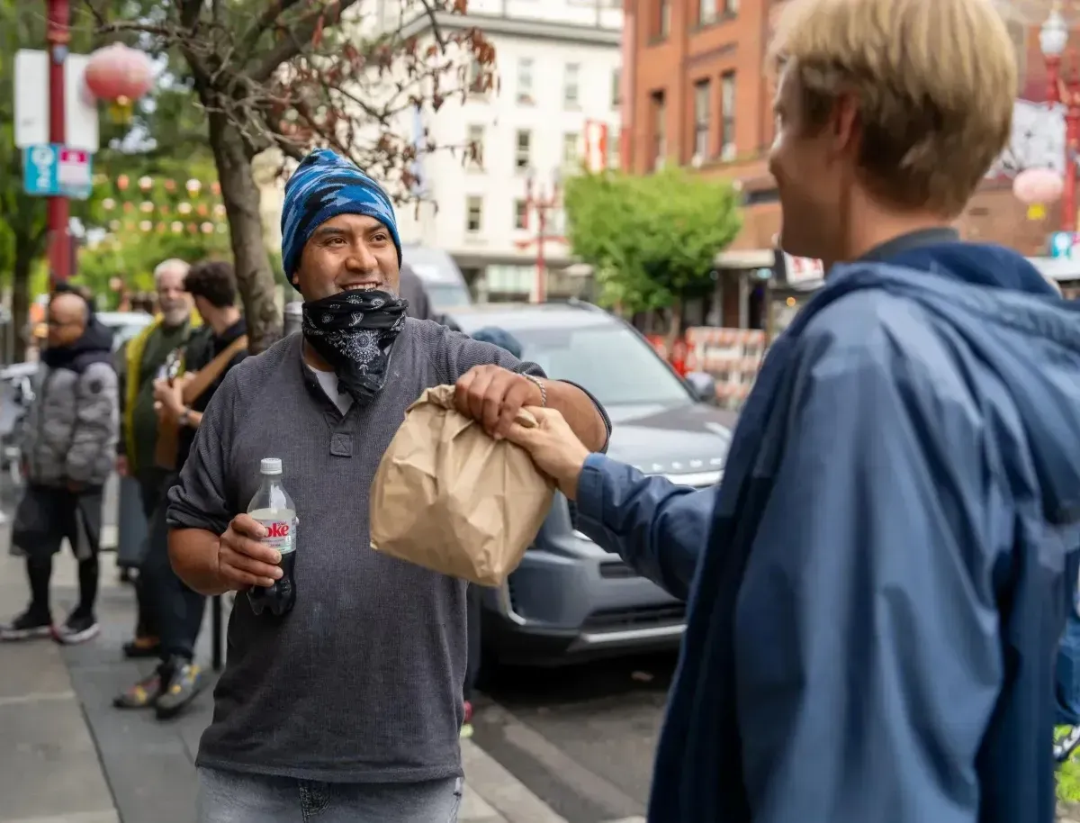 Two people exchanging a brown paper bag on a city street, one holding a drink cup, with parked cars behind.