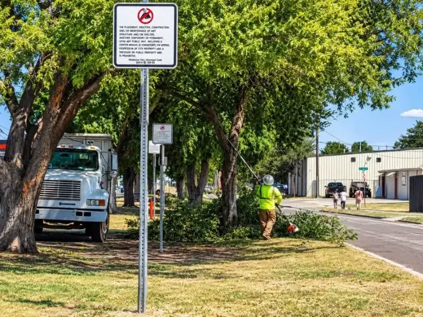 City crew clearing brush under trees beside a no-camping sign on public property in Oklahoma City