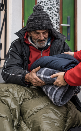 Person wrapped in a sleeping bag being assisted at a doorway, wearing a hooded jacket and red shirt