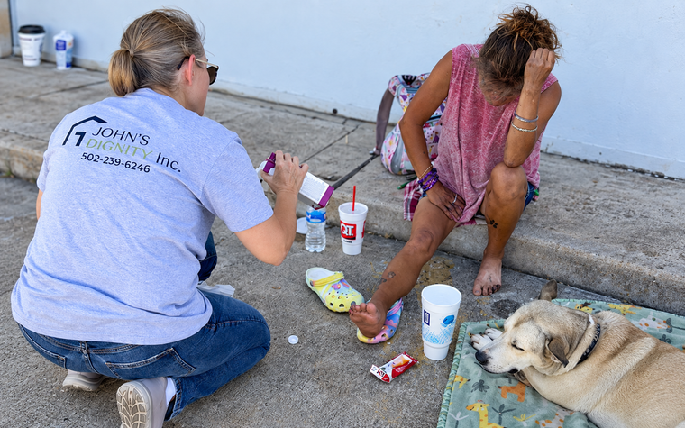 Two people sit on a porch sharing food with a small dog lying beside them.