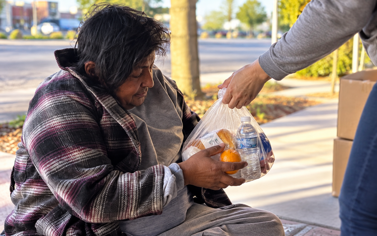 Person seated outdoors receives a bag of food from another person’s outstretched hand.