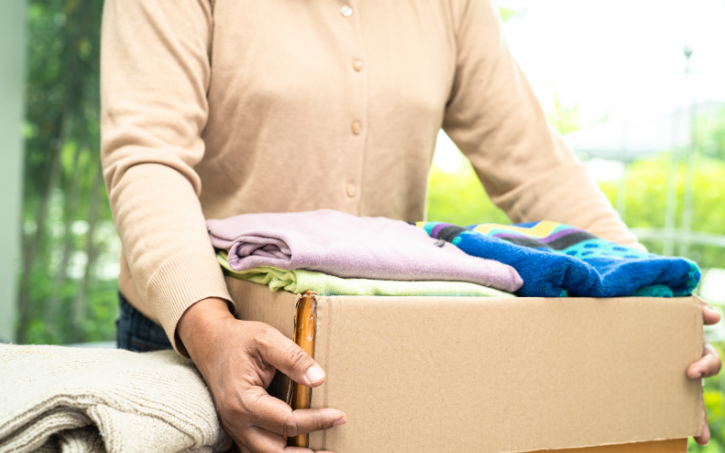 Person holding a cardboard box filled with folded clothes, ready for donation. Sunlight filters through a window in the background.