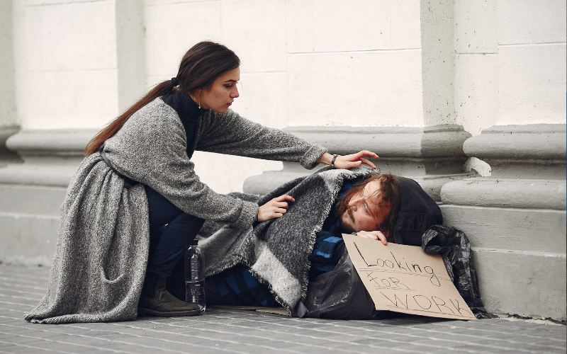 A woman comforts a homeless man sleeping on the street, with a "Looking for Work" sign beside him, showing compassion and need.