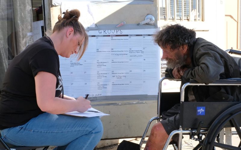 A woman helps a man in a wheelchair, filling out forms beside a schedule board, highlighting community support and care.