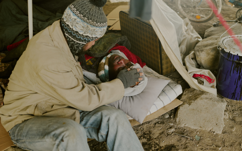 A man in a knit hat tends to another person lying on blankets in a makeshift shelter, conveying care and compassion.