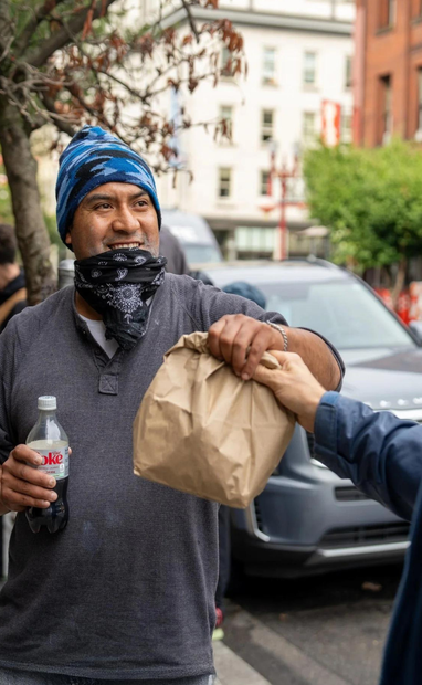 Person in blue beanie and black mask receives a brown paper bag and holds a bottle on a city street.