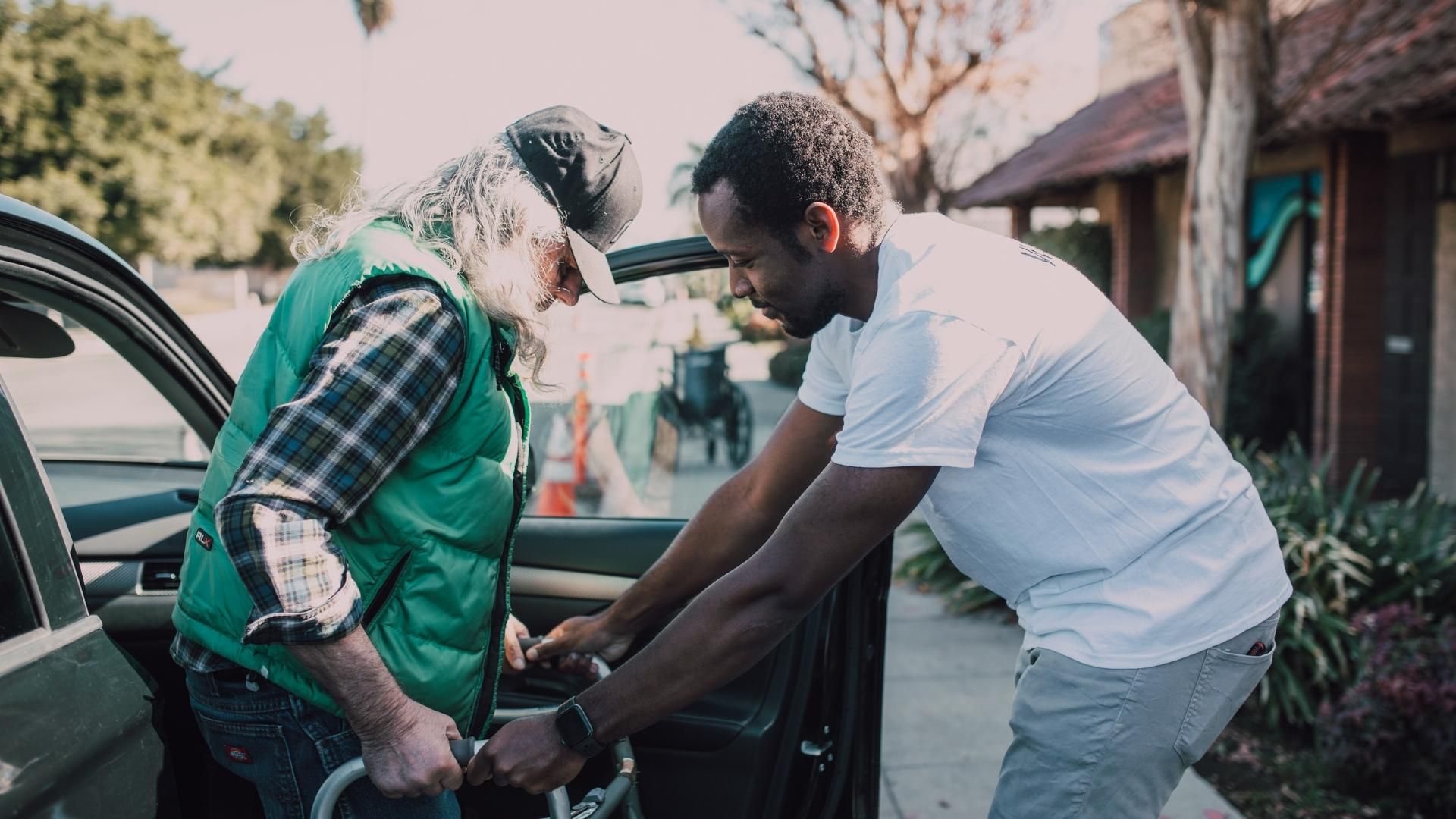 A man assists an elderly person with a walker out of a car, showcasing kindness and support in a sunny outdoor setting.