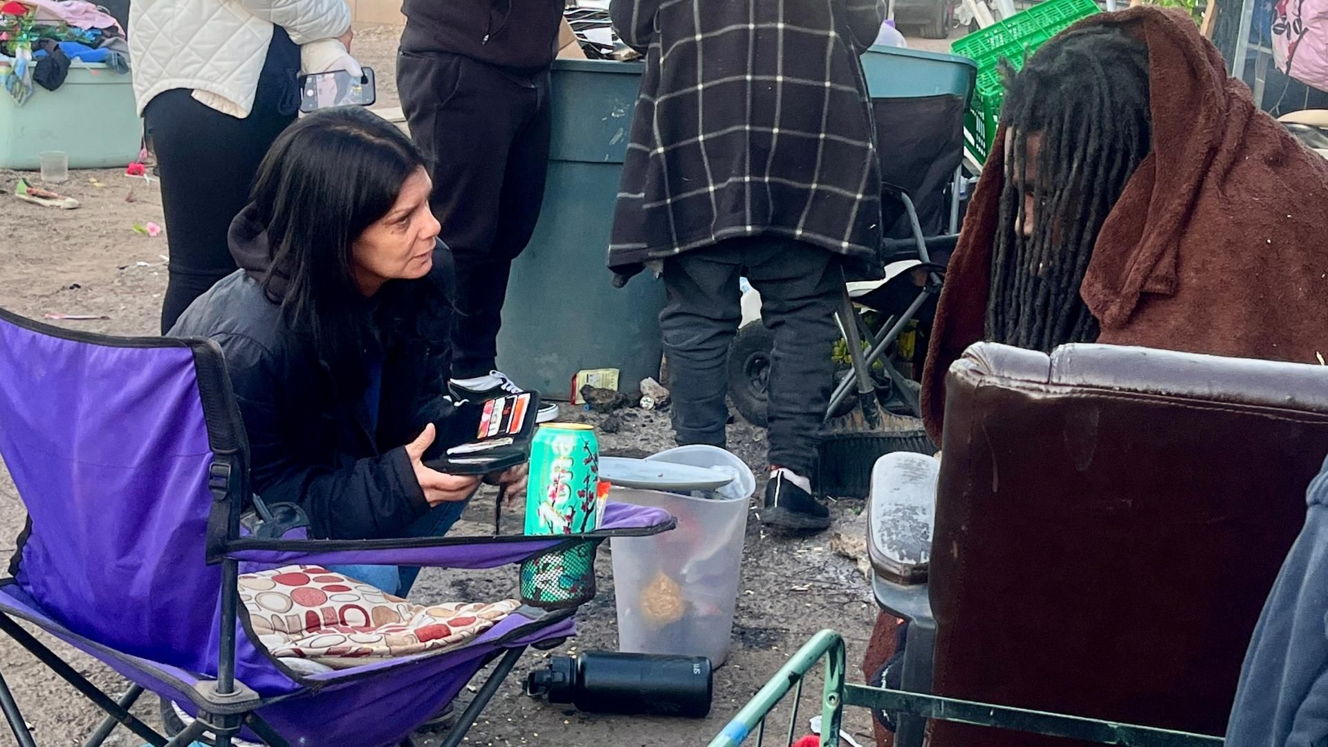A woman in a purple chair talks to a man wrapped in a brown blanket, surrounded by clutter and a can of Arizona tea.