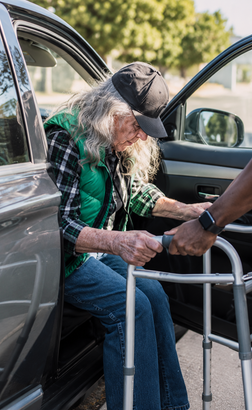 Person seated in a walker at an open car door, being assisted by another person outdoors