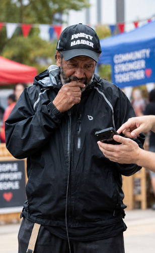 Man in black rain jacket and cap eating at an outdoor community event, with blurred banners behind him