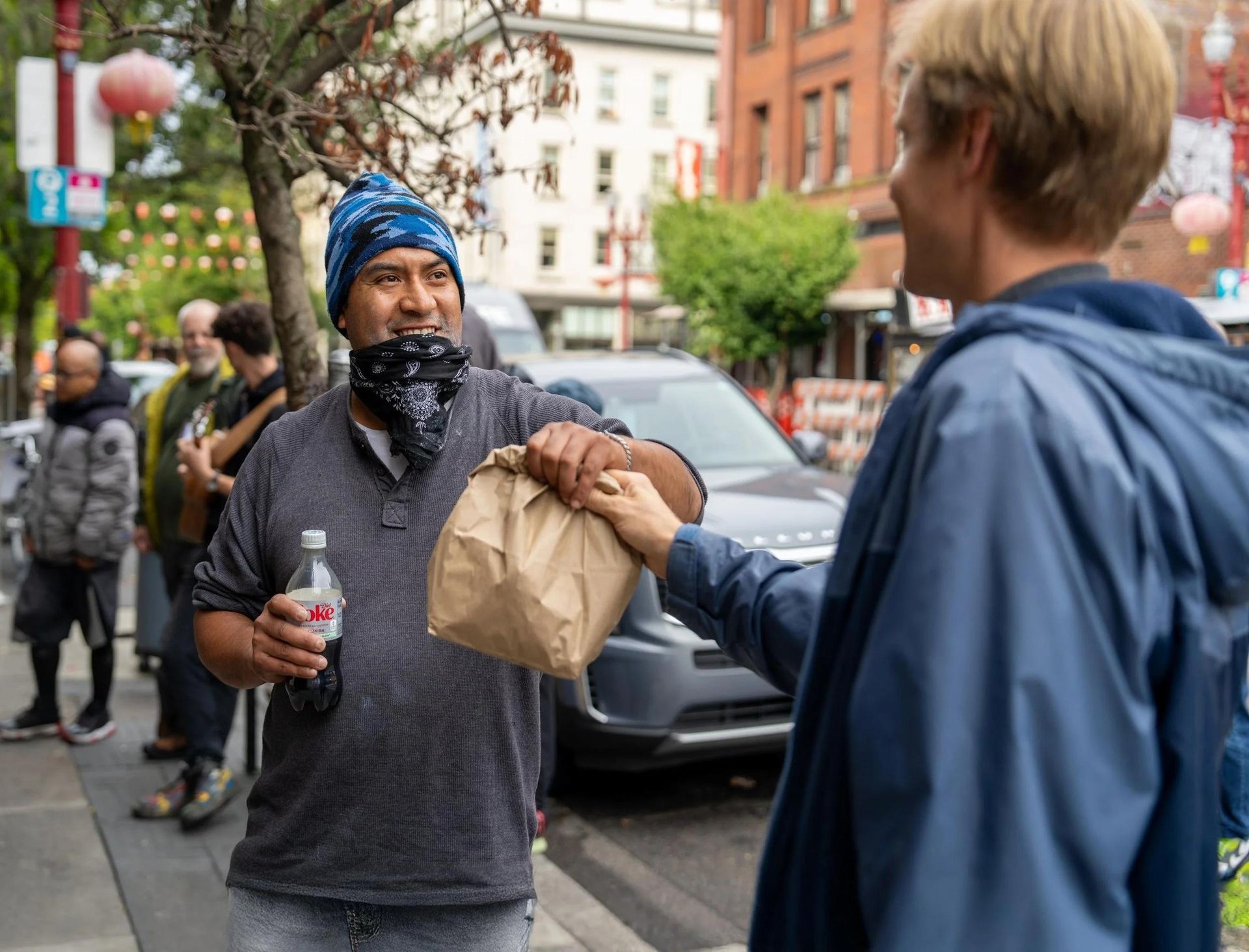 Two people exchanging a brown paper bag on a city street, one holding a drink cup, with parked cars behind.