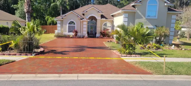 Single-story suburban house with a red driveway, front yard shrubs, and a yellow caution tape barrier.