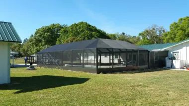 Black mesh-enclosed patio on a grassy lawn beside white buildings and trees under a blue sky