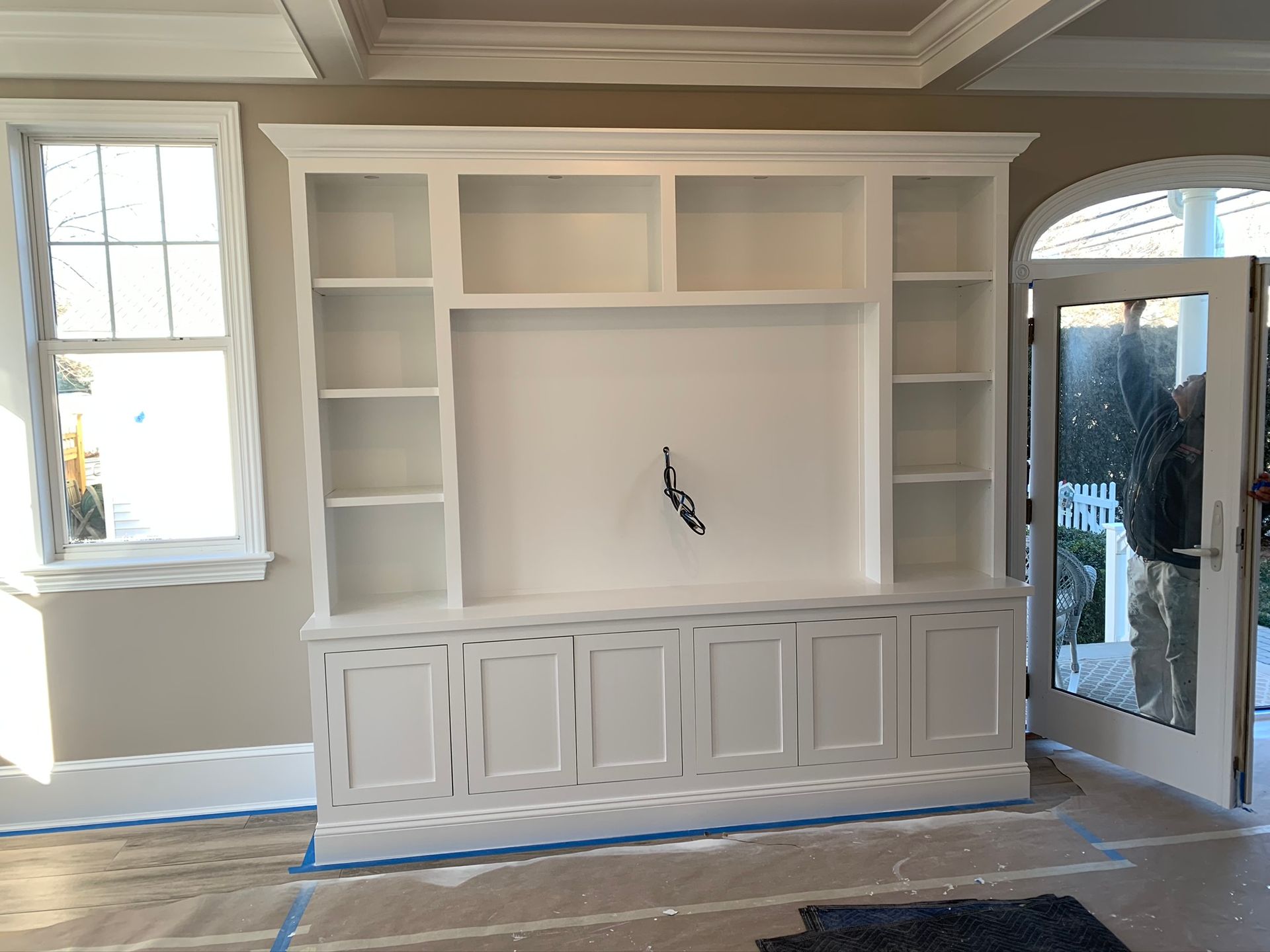 White built-in entertainment center with cabinets and shelves in a bright room, next to a window and glass door.