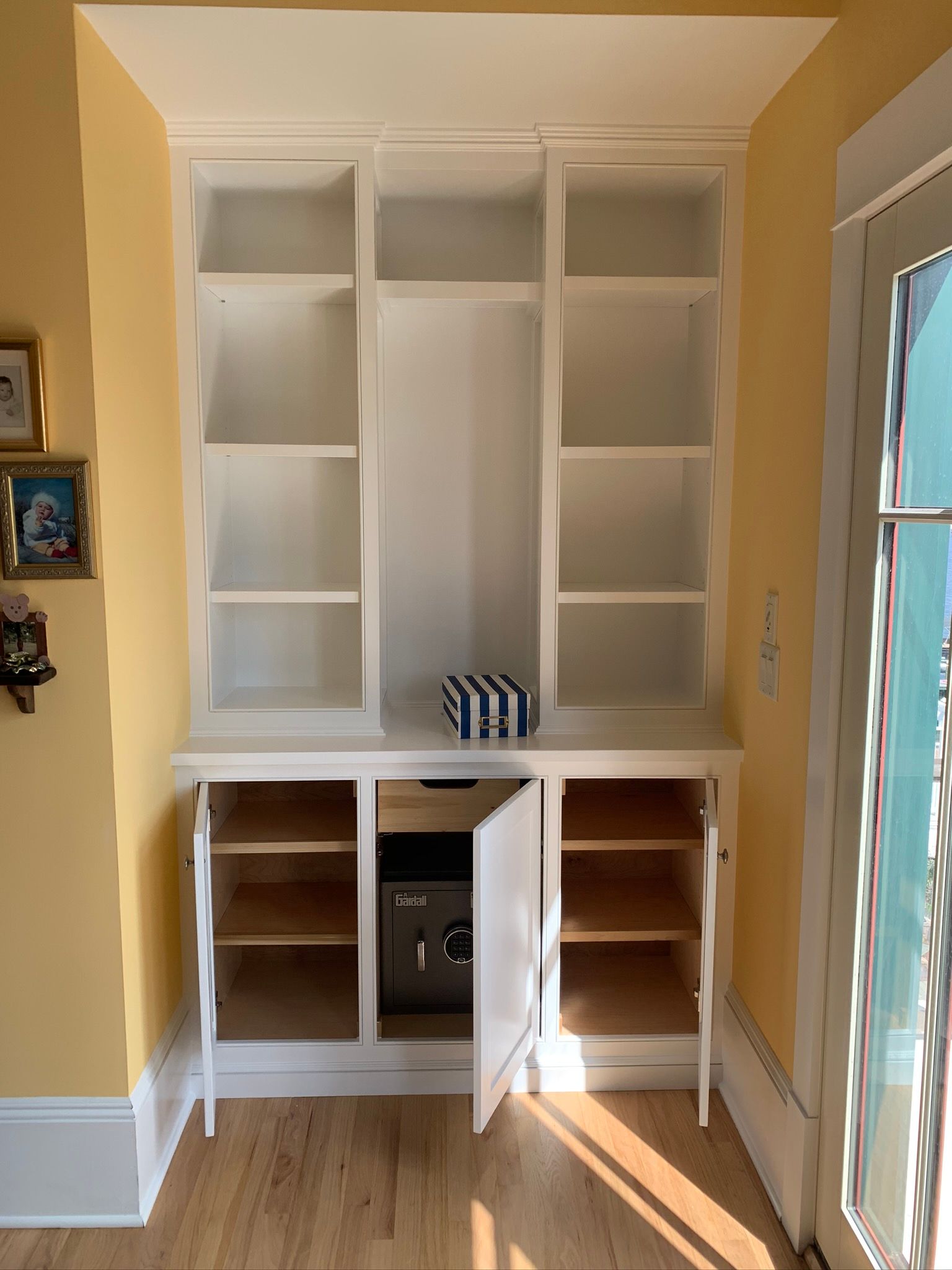 Built-in white shelving and cabinet nook with open doors in a sunlit room, beside a glass door.