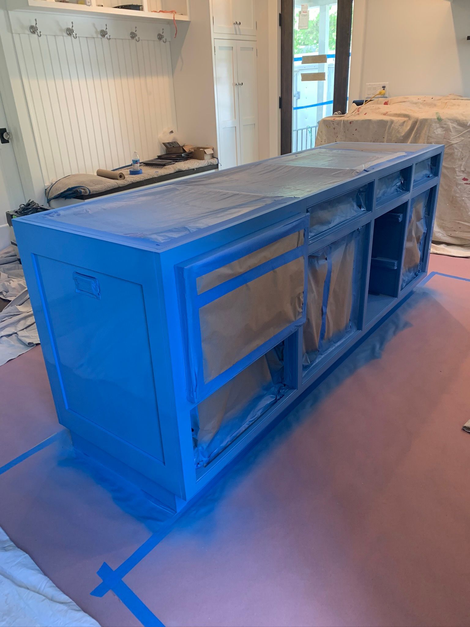 Blue-taped kitchen island base in a room under renovation with cabinets and plastic-covered furniture in the background