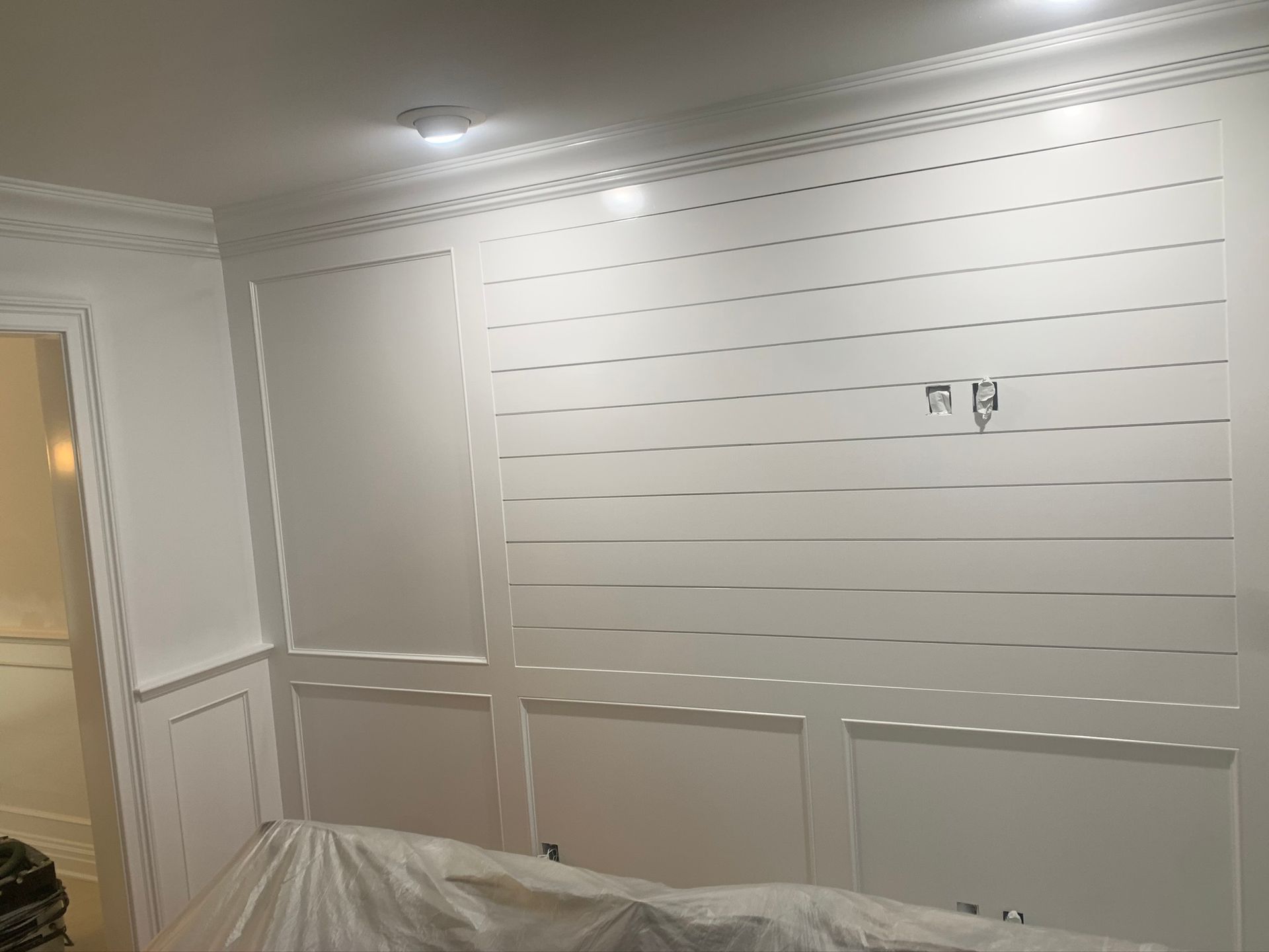 White paneled interior wall with molding and overhead lights, room under renovation with drop cloths on the floor