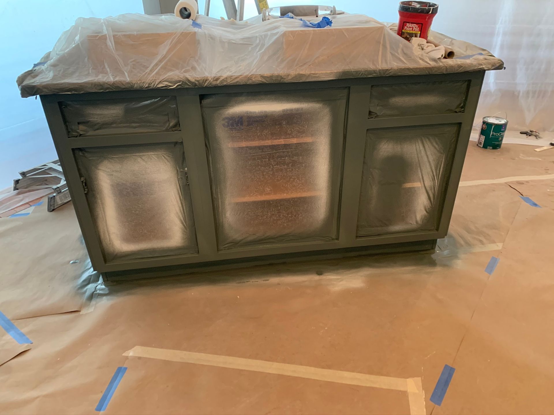 Kitchen island cabinets taped off for painting, with plastic-covered countertop in a room under renovation