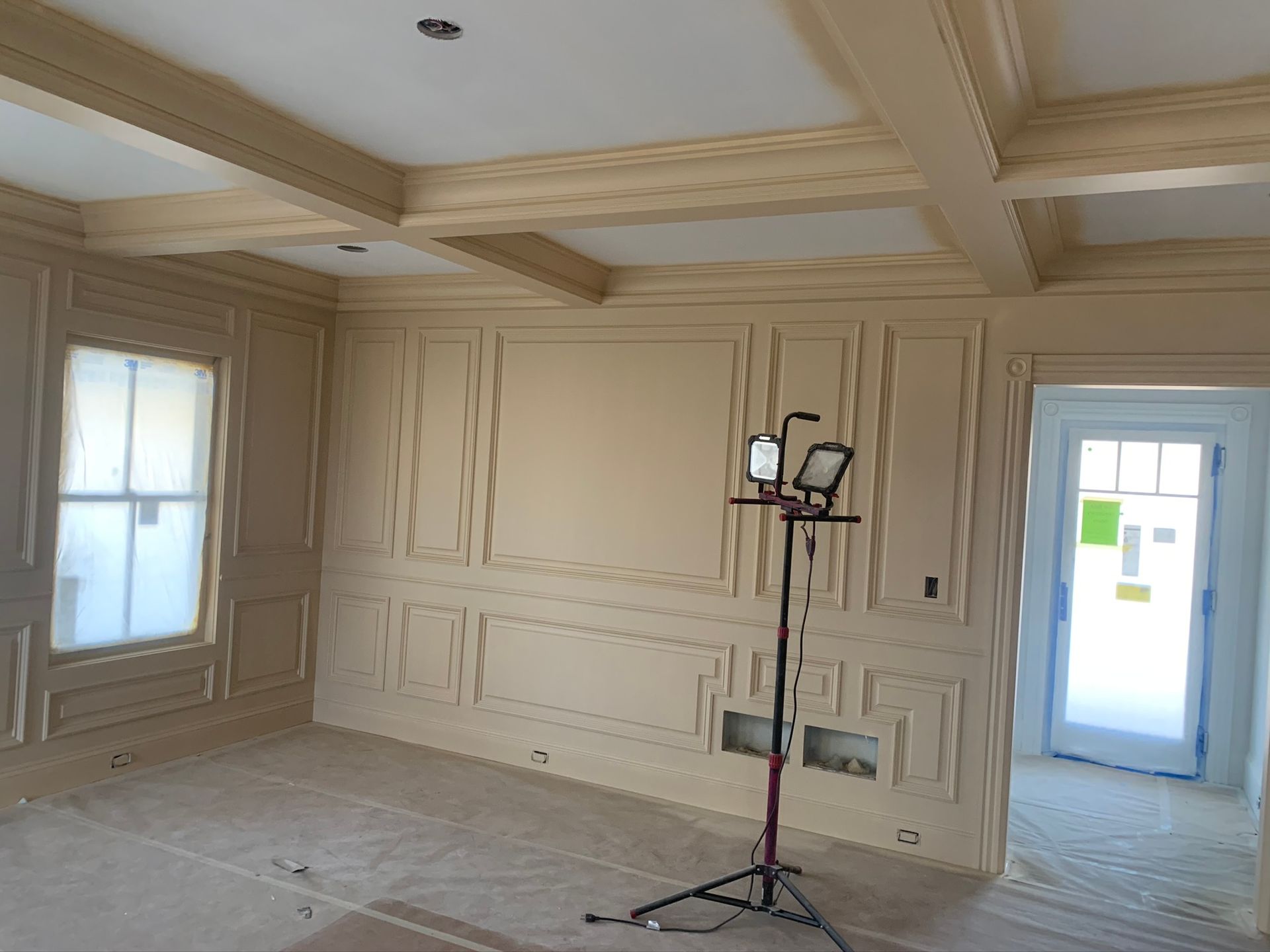 Empty beige-paneled room with large windows and a bright doorway, lit by a tripod work light.