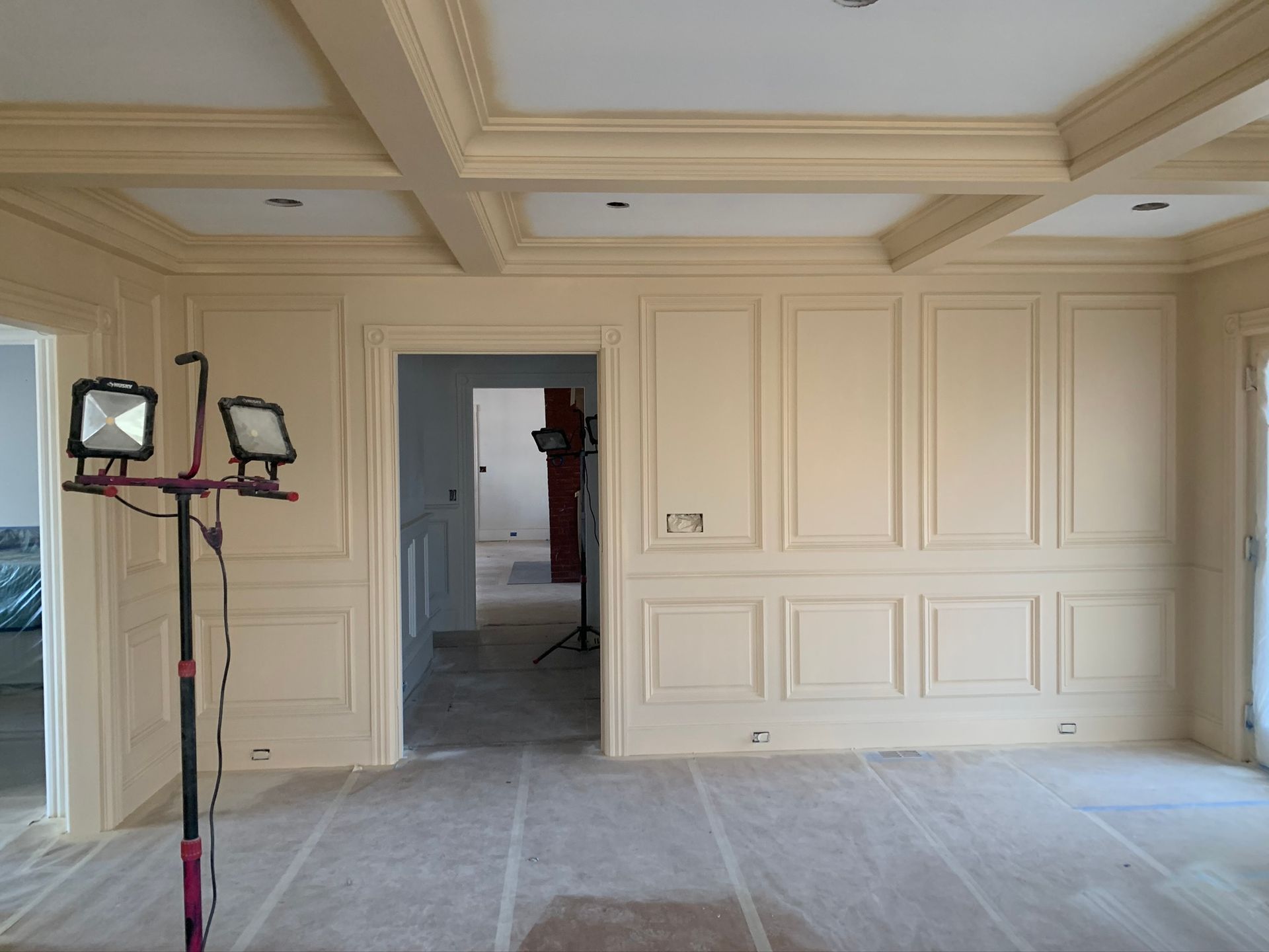 Empty cream-paneled room with coffered ceiling, unfinished floors, and work lights on stands.