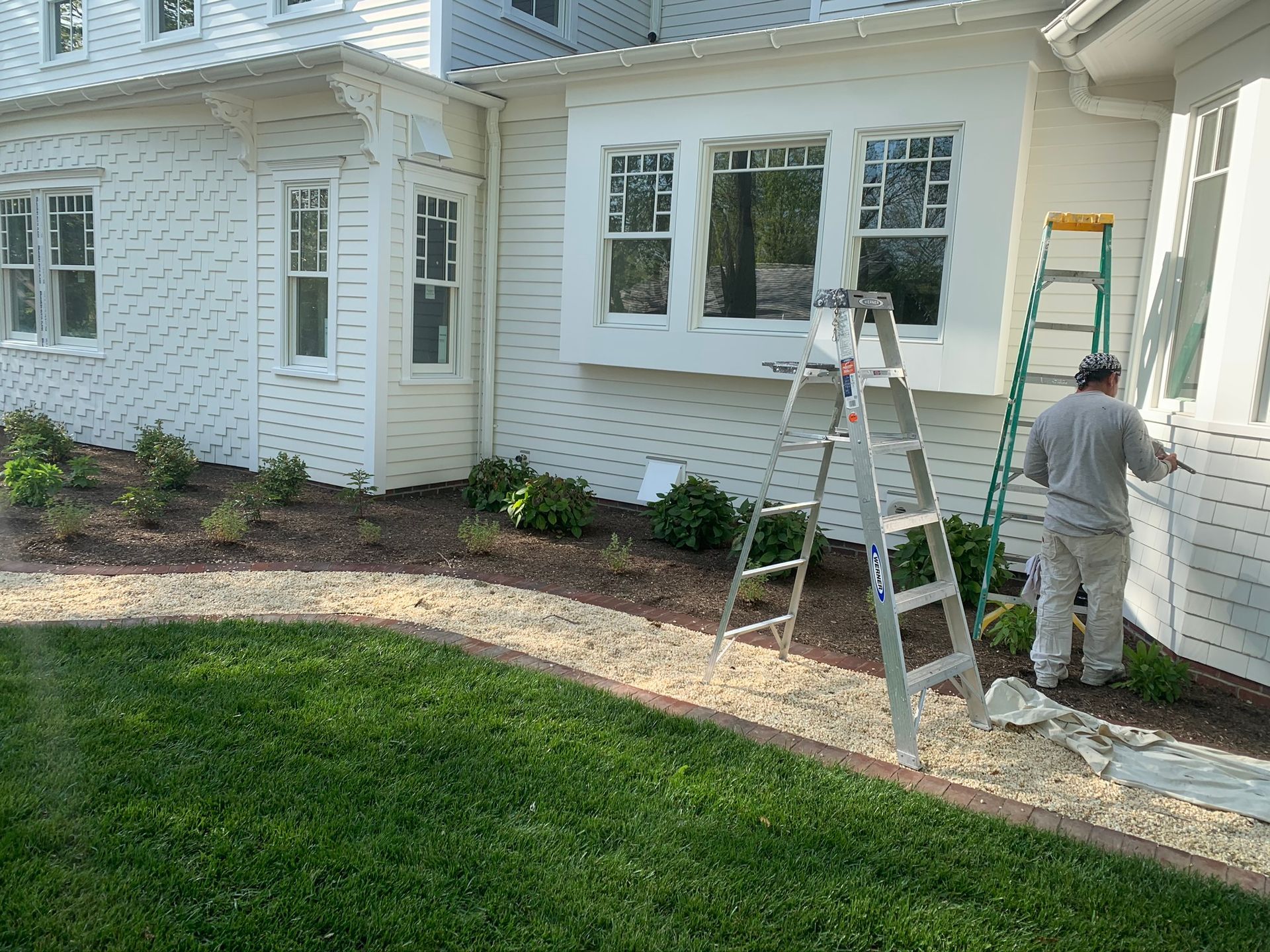 Workers painting a white house exterior beside ladders and a landscaped walkway