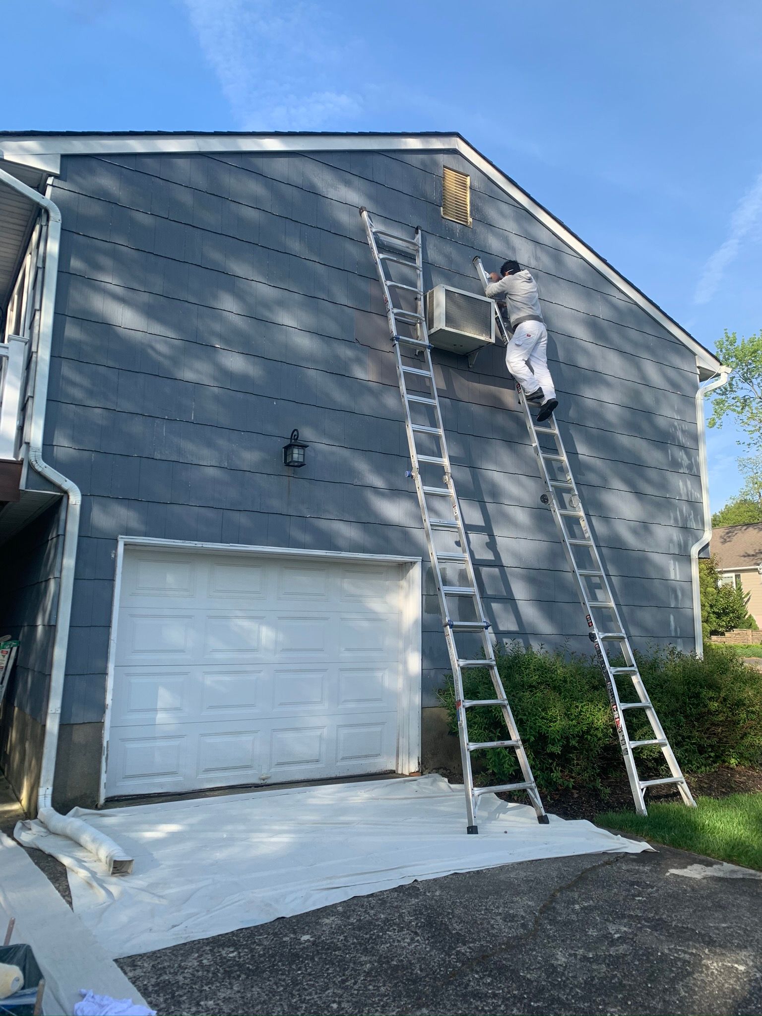 Worker on a ladder painting gray exterior siding on a house beside a garage.