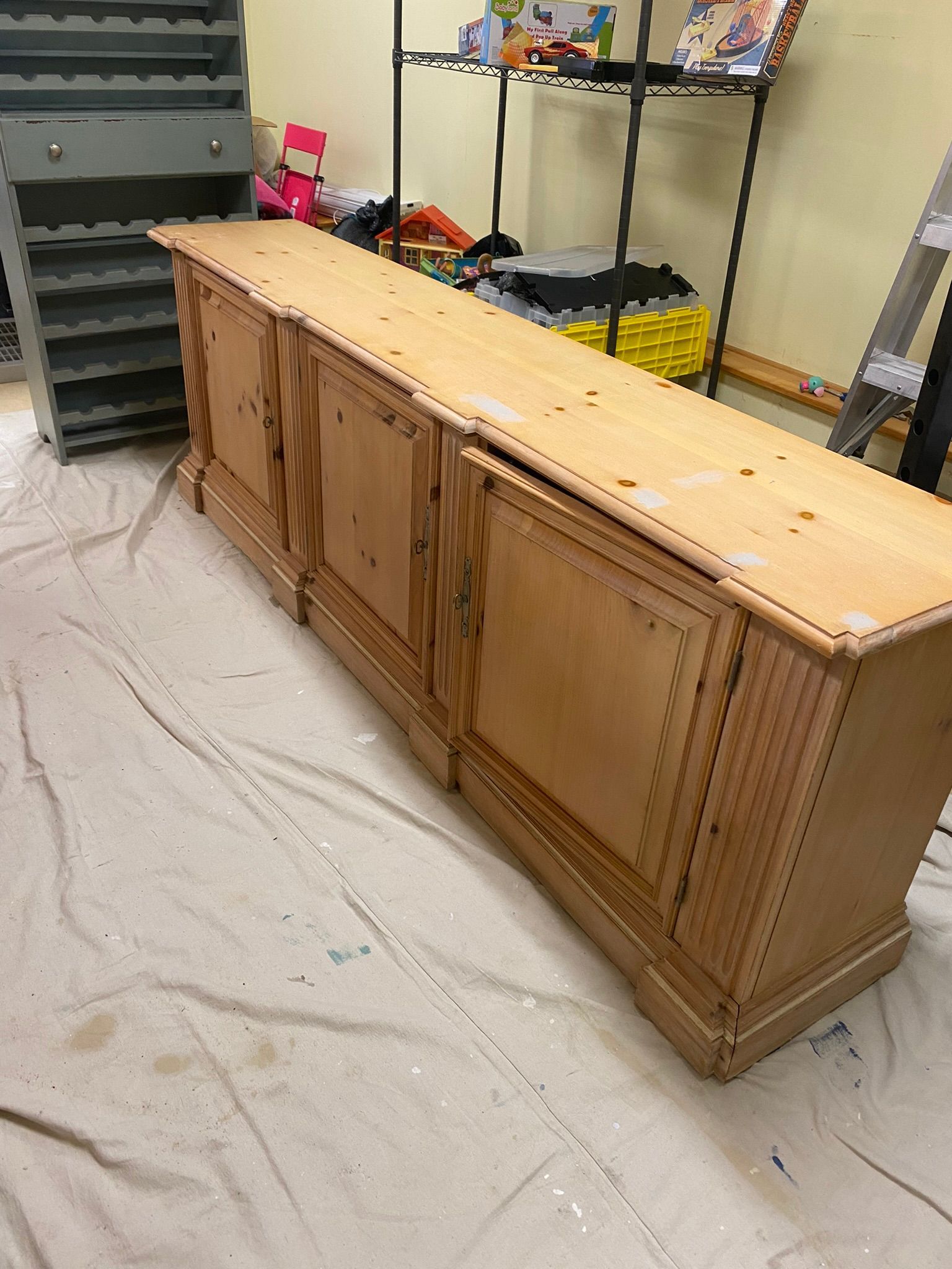 Long wooden sideboard on a drop cloth in a workshop or garage, with shelves and storage bins in the background