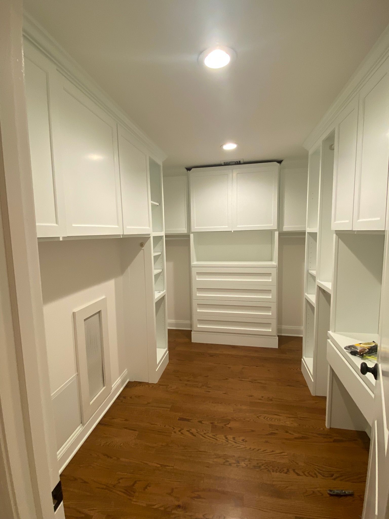 Narrow white walk-in closet with built-in shelves, drawers, and warm wood flooring under recessed lights