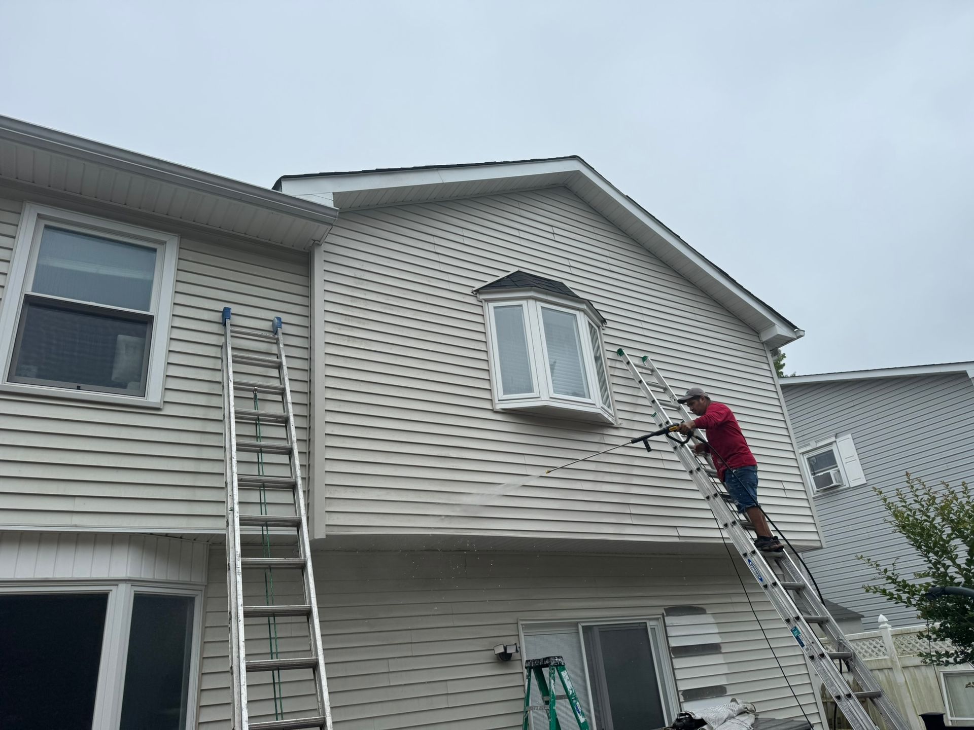 Worker on a ladder painting the siding of a beige house under an overcast sky