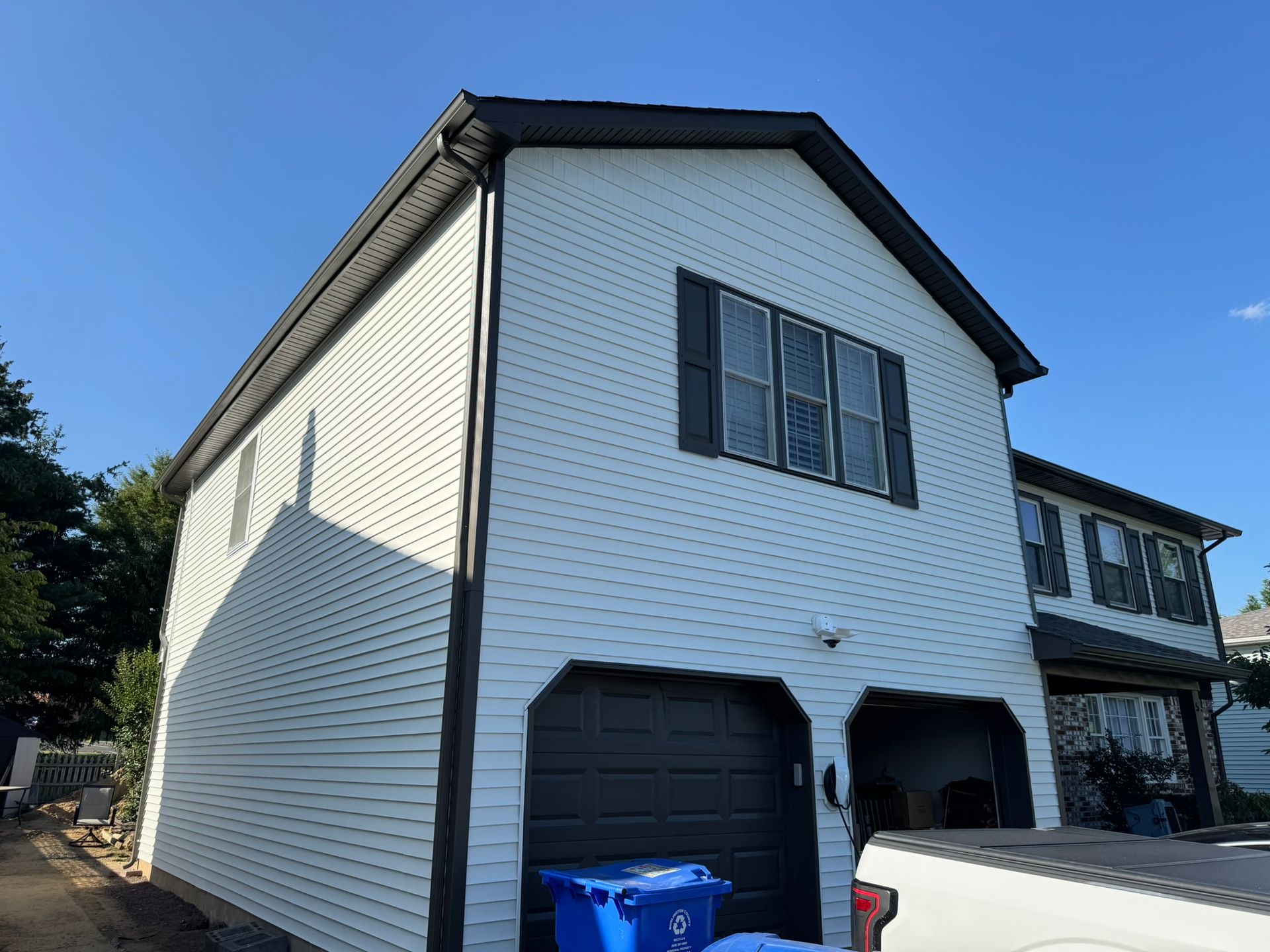 White two-story house with black garage doors and blue recycling bins under a clear blue sky