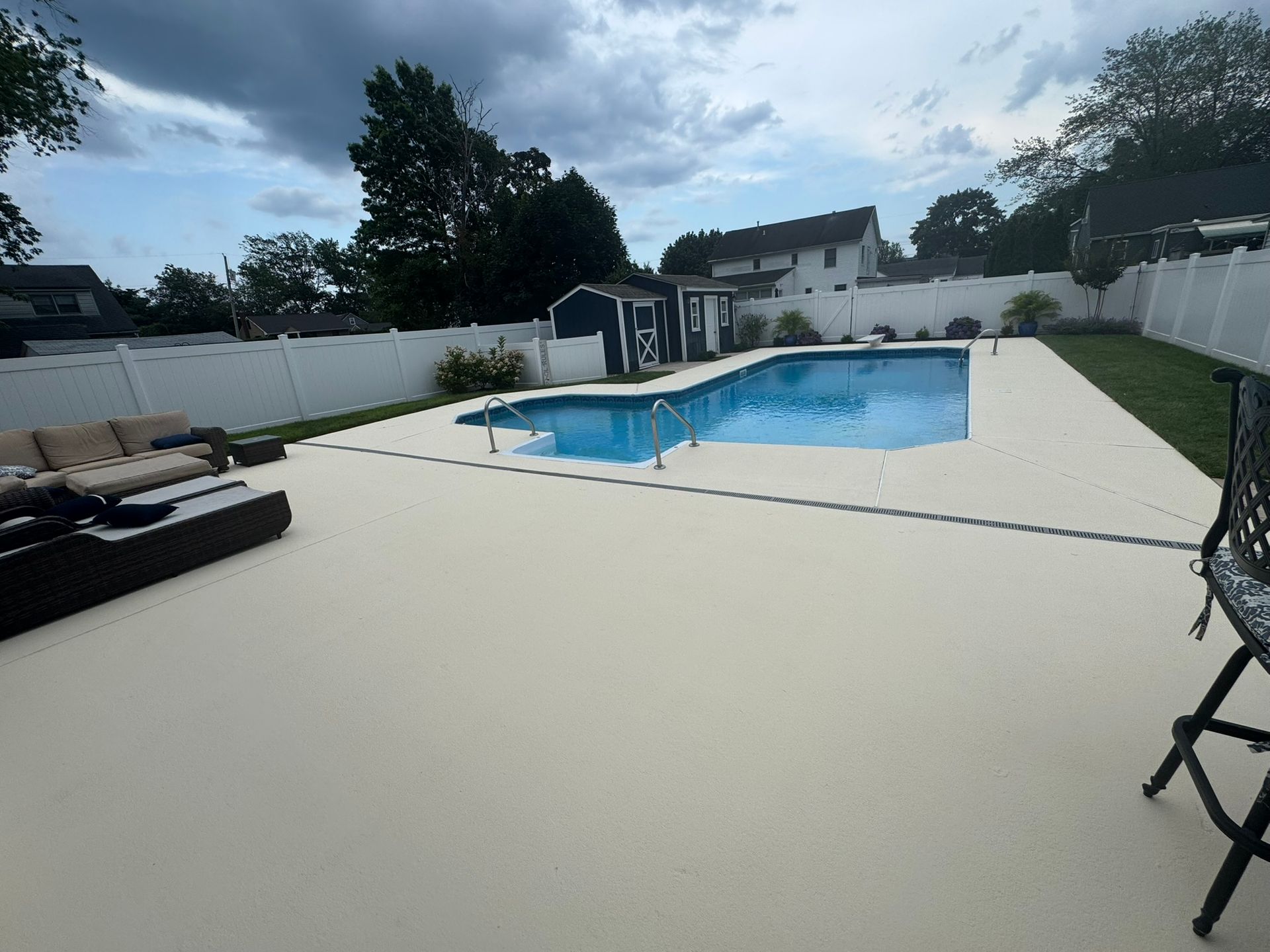 Backyard pool with white patio, lounge chairs, and fenced yard under cloudy sky